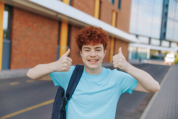Redhead teen boy smiles and gives thumbs-up outside modern school