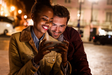 Smiling couple using mobile phone at night in city outdoors