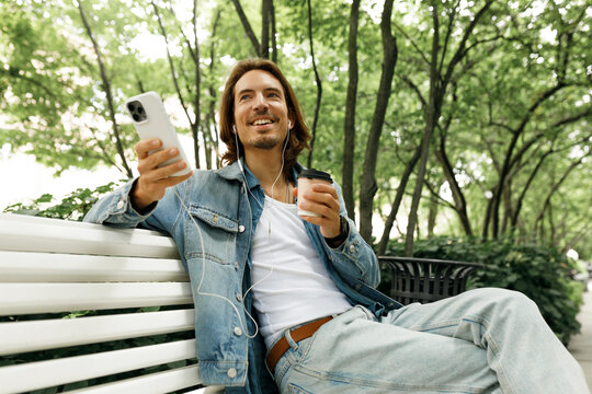 Low angle view of smiling artist having rest relaxing in city park, sitting on bench leaning his back against it and listening to favorite music, enjoying coffee and good summer evening