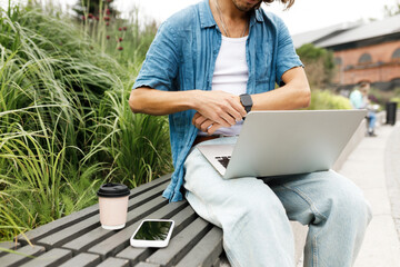 Outdoor image of unrecognizable man freelancer in stylish denim clothes working online on laptop sitting on bench in public park, checking time on his wristwatch, deciding to have coffee break