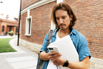 Good-looking adult IT-specialist freelancer with long hair holding his laptop and using application on his smartphone, reading work chat on his way to have working day outside in park or cafe