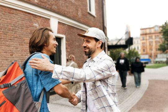 Side view outdoor image of two smiling caucasian men in casual clothes shaking hands and giving each other hugs after sudden meeting on city street, being hurry for work, having small talk