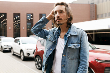 Choosing car concept. Stylish caucasian man with long hair in denim shirt scratching his head...