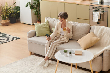 Senior woman enjoying a cozy moment at home with a drink and smartphone in hand