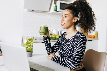 Side view of slim african american woman starting her day with detox smoothie, sitting at kitchen table in front of laptop and drinking homemade green fresh juice