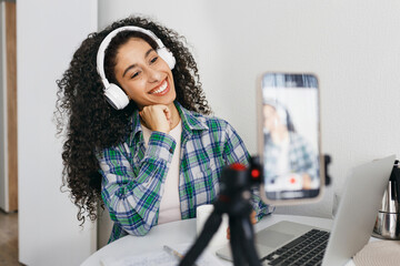 Happy generation z female streamer or blogger of african american ethnicity in wireless headphones holding white mug of coffee while talking to her followers on camera of smartphone on tripod