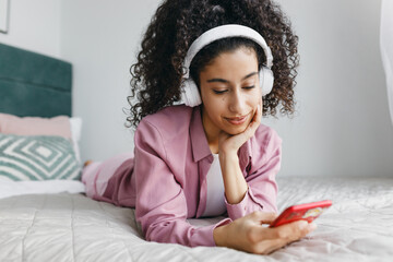 Pretty teen mulatto girl with curly hair relaxing on bed in headphones selecting playlist on her smartphone, enjoying favorite music, listening to new popular songs while chatting in group chat