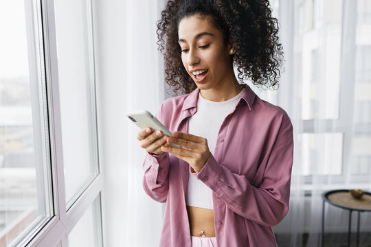 indoor image of black girl in pink shirt browsing social media newsfeed, scrolling comments and chatting online, checking weather forecast using mobile application on her smartphone
