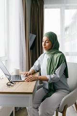 Muslim businesswoman in green hijab using laptop while sitting at wooden desk in cozy home office,...