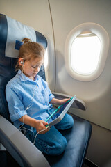 Little girl watching a cartoon on a tablet with headphones during an airplane flight. Digital entertainment, child focus, and quiet family travel experience.
