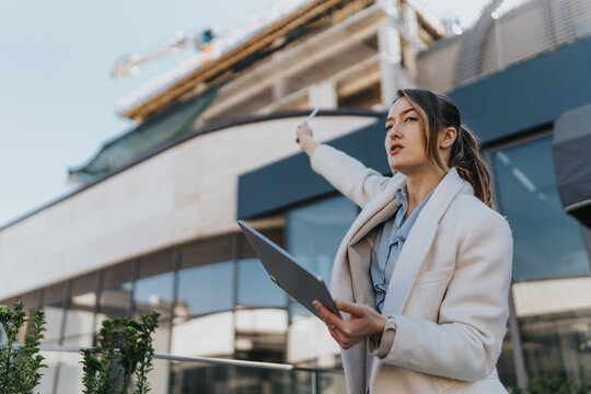 A confident businesswoman evaluates a construction site, holding a tablet while pointing towards the building.