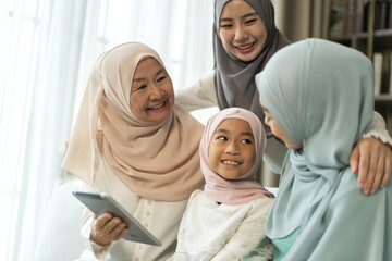 Muslim family four generations sitting on sofa enjoying tablet, joyful moment with grandmother,...