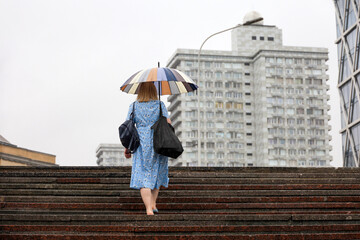 Woman in summer dress with umbrella walking up the steps on city buildings background. Heavy rain...