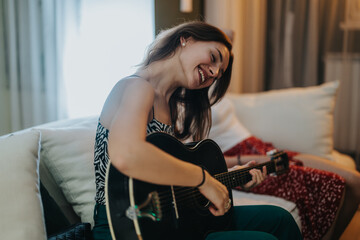 A cheerful woman smiles as she plays an acoustic guitar while sitting on a sofa, enjoying a relaxed...