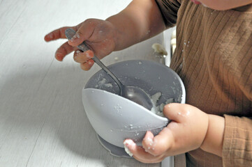 Toddler learning to eat independently with a spoon, holding a bowl with food. Messy eating process....