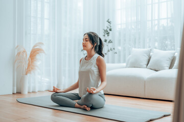 Young Asian woman meditating in lotus position at home, sitting on the floor in fitness clothing....