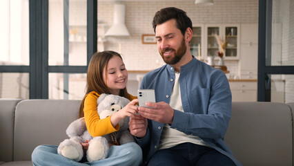 Naklejka na ściany i meble Smiling Caucasian child little girl dad father man touching screen mobile phone smartphone speaking family at home e-commerce glad kid daughter online shopping together parent male Internet cyberspace