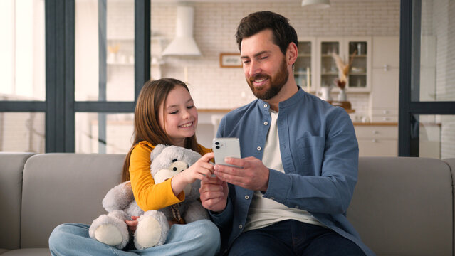 Smiling Caucasian child little girl dad father man touching screen mobile phone smartphone speaking family at home e-commerce glad kid daughter online shopping together parent male Internet cyberspace