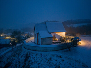 Aerial view of a church warmly illuminated against the cold, snowy landscape, its red accents standing out against the dominant blue, ÄŒerÃ­n, BanskÃ¡ Bystrica Region, Slovakia.