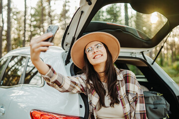 Young caucasian woman taking self portrait in scenic forest while sitting in car trunk
