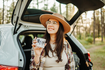 Young caucasian woman drinking water from metal cup in the forest while sitting on car trunk