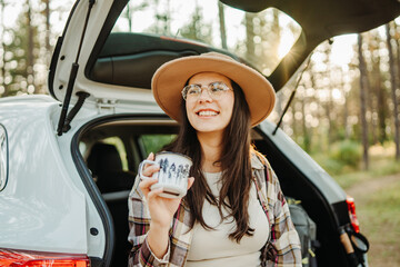Young caucasian woman drinking water from metal cup in the forest while sitting on car trunk
