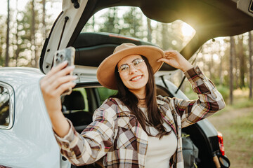 Young caucasian woman taking self portrait in scenic forest while sitting in car trunk