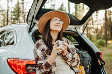 Young caucasian woman drinking water from metal cup in the forest while sitting on car trunk