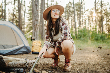 Young caucasian woman preparing tent and campfire in the scenic forest during golden hour