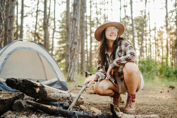 Young caucasian woman preparing tent and campfire in the scenic forest during golden hour