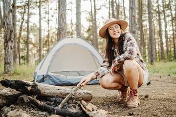Young caucasian woman preparing tent and campfire in the scenic forest during golden hour