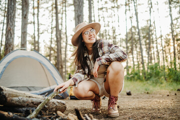 Young caucasian woman preparing tent and campfire in the scenic forest during golden hour