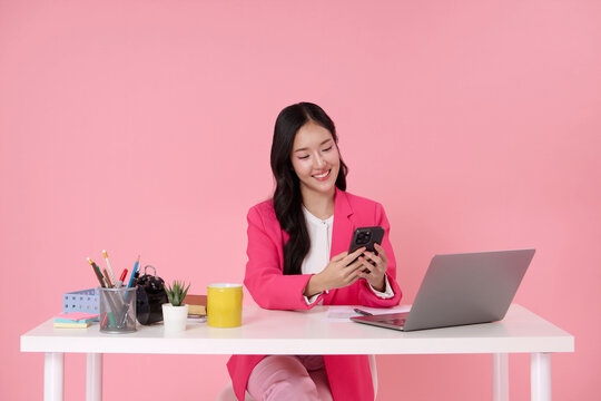 smiling Asian businesswoman in a pink blazer sit at her desk happy engaged with her smartphone with laptop and office supplies around her on pink background. - Powered by Adobe