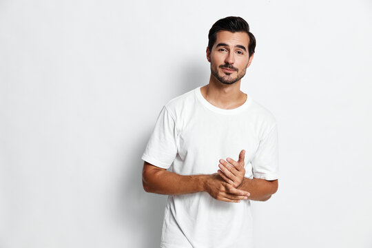 Full body thoughtful smiling young man wearing a white t-shirt looking at the camera, standing with hands together against a plain white background. People lifestyle concept.