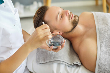 Cosmetologist applying moisturizing mask with brush to a young relaxed smiling man in spa salon....