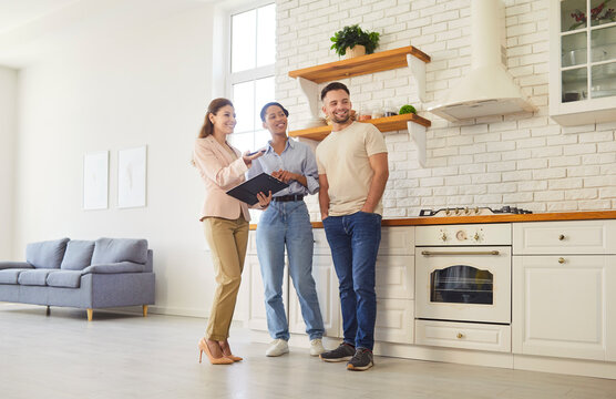 Female broker giving tour for family couple, showing spacious house, demonstrating kitchen. Husband and wife planning to buy new home, consulting expert. Purchasing and mortgage concept