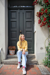 Young woman sitting on doorstep under blooming rose bush in front of vintage house facade