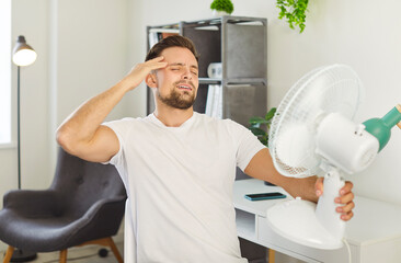 Portrait of young unhappy man employee standing on workplace and suffering from summer heat in front of fan. Male person using electric fan during heatwave in modern office indoors.