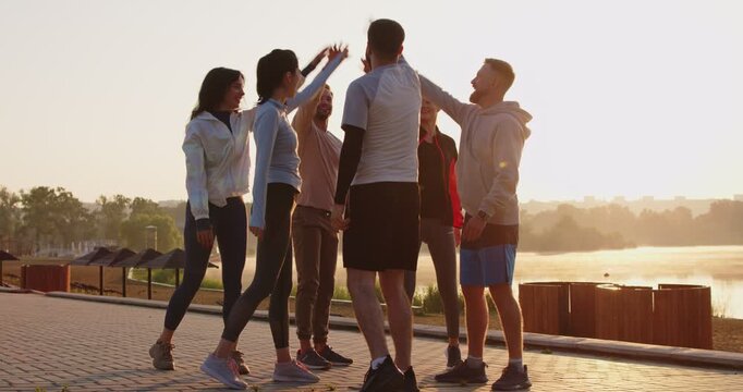 Group of positive people stands in circle, holding hands together, celebrating success during outdoor workout, boosting sport fitness, uniting in training joy in a shared open setting.
