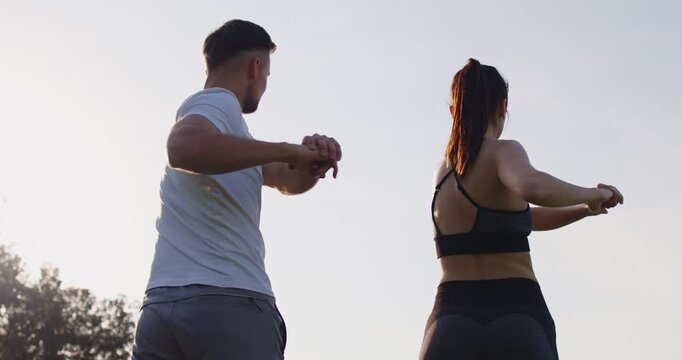 Young couple exercising outdoors. From behind 2 people in fitness sportswear standing against sky background in park doing trunk rotations for vitality, energy, core strength and flexibility