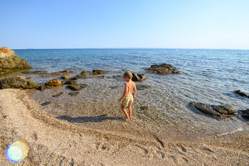 A boy walking in shallow water among pebbles on a wild beach. Quiet moment of exploration, nature connection, and carefree childhood at the seaside.