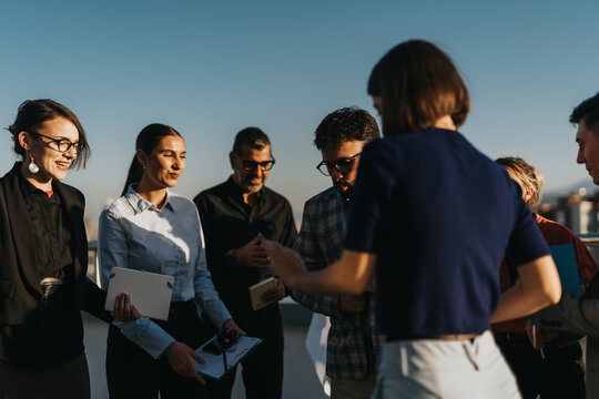 A diverse group of business people relax and socialize on a high-rise balcony at sunset, enjoying music and conversation. This setting portrays teamwork, relaxation, and multicultural cooperation.