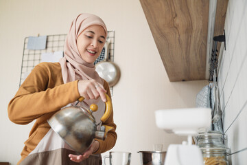 Asian Muslim woman wearing a headscarf pours water from a kettle in a cozy kitchen setting,...