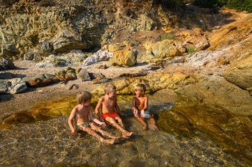 Little boys playing, laughing, and swimming among rocks in the sea on a wild beach. Joyful summer moment full of energy, freedom, and childhood adventure in nature.