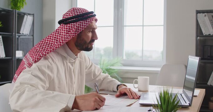 Young Arab businessman sitting at the desk waving hand having video call on a laptop in office. Confident Muslim man in traditional dishdasha dress working online having webinar. 4k video.
