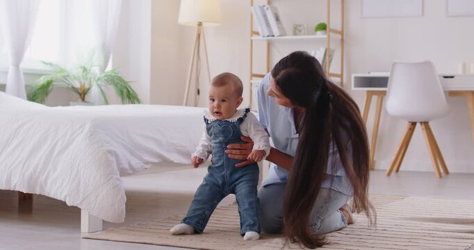 Happy cheerful mother sitting with her infant little cute child daughter supporting her in first steps at home. Toddler girl stepping on floor learning to walk with mom help. 4k video. Slow motion.