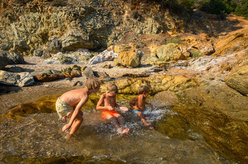 Little boys playing, laughing, and swimming among rocks in the sea on a wild beach. Joyful summer moment full of energy, freedom, and childhood adventure in nature.