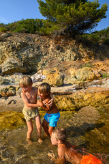 Little boys playing, laughing, and swimming among rocks in the sea on a wild beach. Joyful summer moment full of energy, freedom, and childhood adventure in nature.