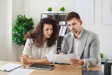 Two young confident smiling business people and company employees in formal clothes working in office at the desk looking through financial documents on workplace discussing work project.