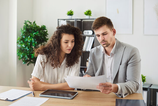 Fototapeta Two young confident smiling business people and company employees in formal clothes working in office at the desk looking through financial documents on workplace discussing work project.